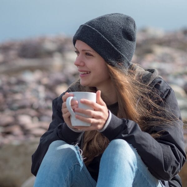 Young woman sitting outside on rocks near the seaside holding a white mug and wearing a grey beanie hat, ripped jeans and a black hoodie.